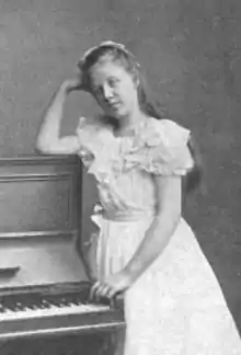 a young white girl with long hair, leaning on a small upright piano, wearing a white frock.