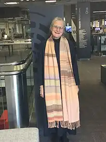 Bridie Lonie stands in the Alexander Turnbull Library. She is smiling and wears a long and colorful woven scarf, over dark clothes.