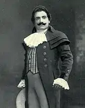 Man, with dark hair and a curling moustache, standing in a posed position. He is wearing a long coat, with lace at the throat and cuffs.