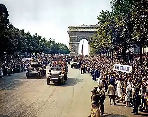 Crowds line the Champs Elysees as tanks and half tracks roll down it. The Arc de Triomphe is in the background. People are holding a sign that reads: "Viva de Gaulle".