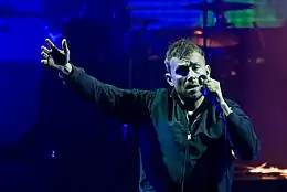 A middle-aged man standing in front of a coloured background with his right arm raised. He is holding a microphone in the opposite hand.