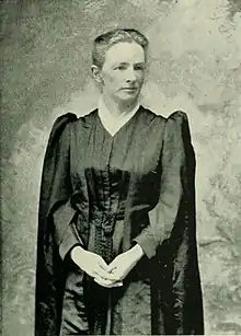 A white woman, standing, in a black clergy robe, with hands folded.