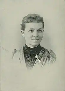 B&W portrait photo of a woman with her hair in an updo, wearing a clergy collage, dark bloues, and light jacket with a white ribbon pinned upon it.