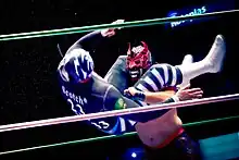 A masked wrestler executing a flying headscissorts, wrapping his legs around the head of his opponent, trying to throw him to the ground.
