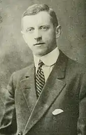 Black and white photo of a man wearing a dark suit jacket, a high-collared white dress shirt, and a striped necktie