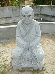 Image of older monk with large eyebrows, sitting, with his hands hidden in his sleeves
