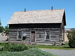 Photograph of a log cabin in a manicured setting with agricultural fields and buildings in the far background
