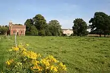 A grassy field, its uneaven surface barely discernable, is bright green under a blue summer's sky. In the foreground are the bright yellow flowers of Rattle. A small brick chapel stands in the rear left corner of the field, and beyond that are a row of mature oaks. A gap in the row of trees gives a view of the frontage of Stainfield Hall