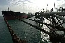 A large red and black tanker ship floats in calm dark waters, tightly moored by ropes to a long steel truss dock, while large pipes pump oil out to the ship