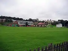 A cricket pavilion joined to a more modern building behind green playing fields, with the sight screens of a cricket pitch to the right